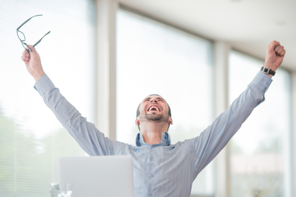 Smiling businessman sitting at desk behind laptop raises arms in the air in a cheer