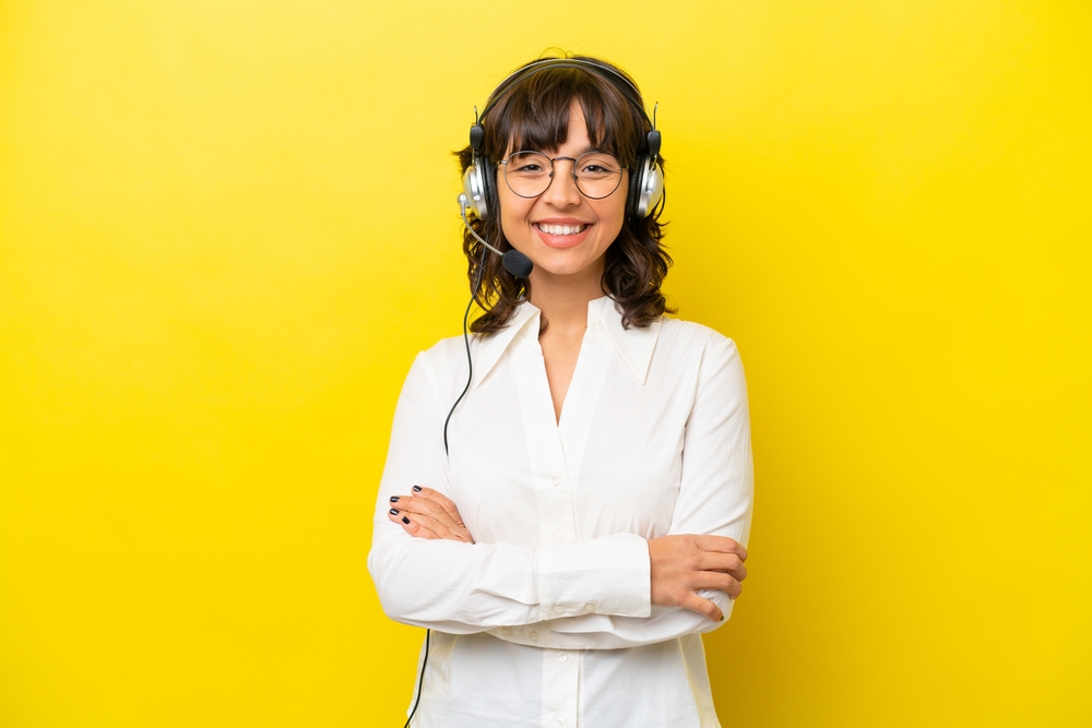 Latinx woman with headset on, smiling, arms crossed, yellow background